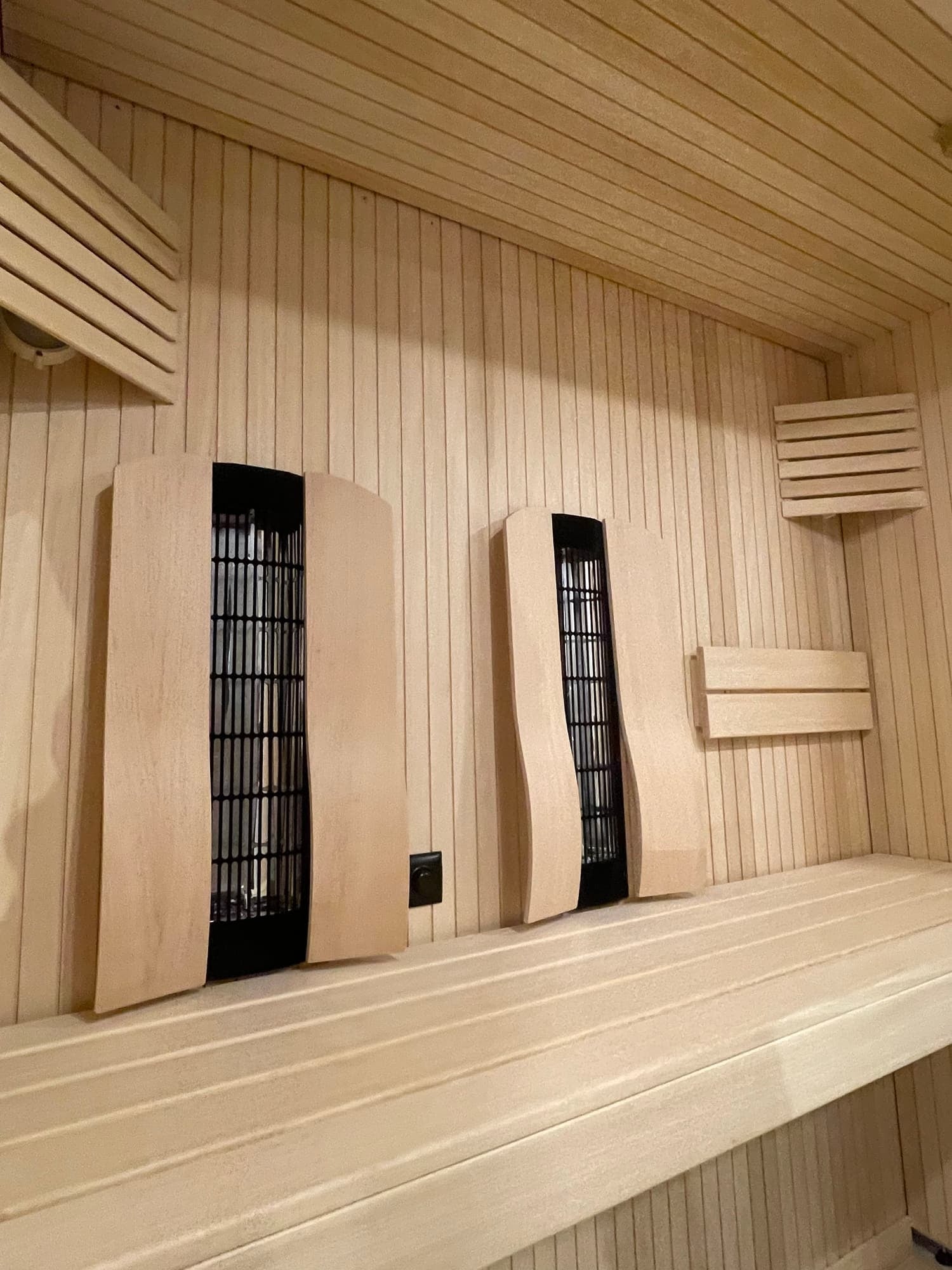 Inside a wooden sauna with two black vent covers on the wall and wooden slats on the walls and ceiling.