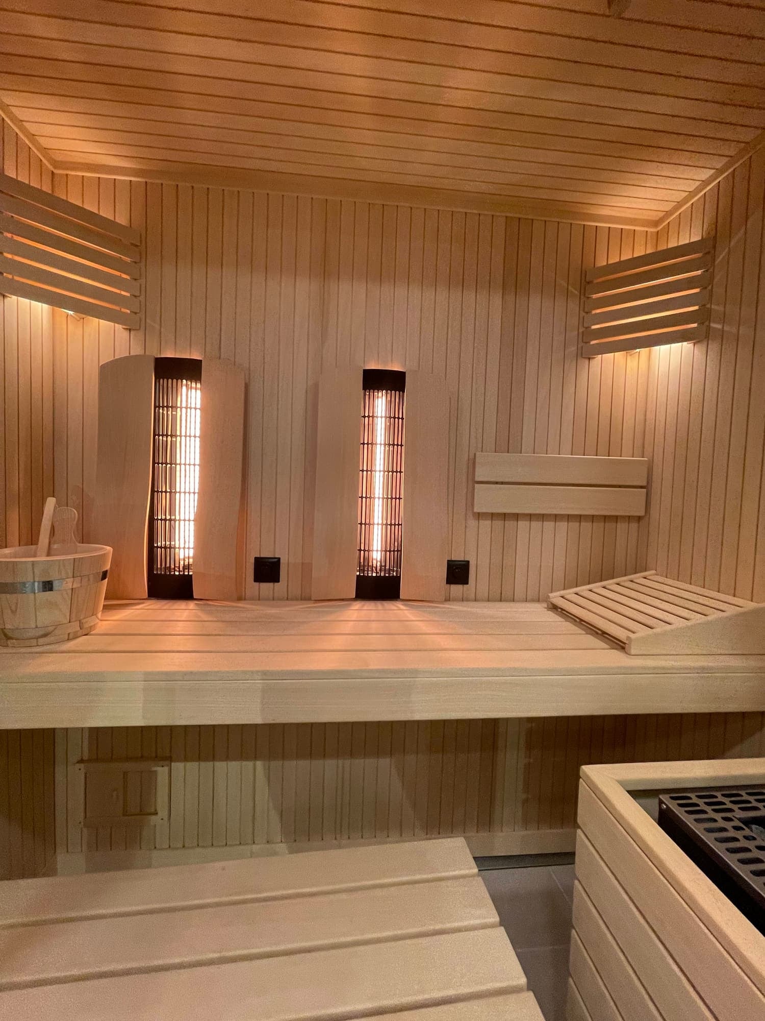 Wood-paneled sauna interior with infrared heaters, a bucket, and wooden benches.