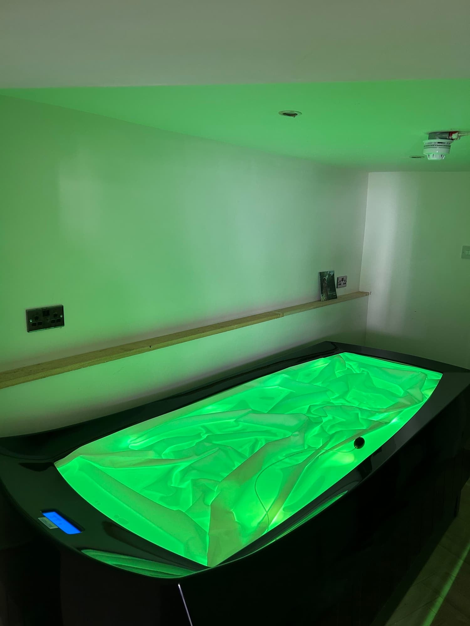 A room with green lighting, featuring a large, rectangular hot tub with a glowing green interior, a wooden shelf along the wall, and electrical outlets.