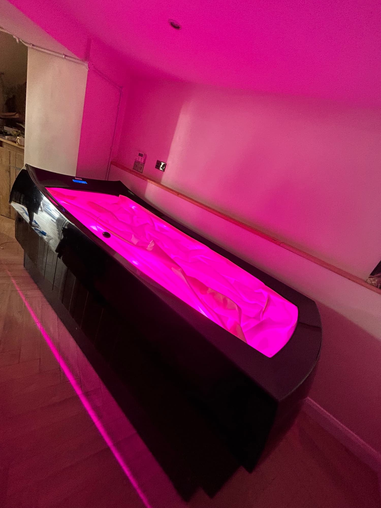 A rectangular sauna with pink lighting inside, placed in a room with pink walls and wooden flooring.