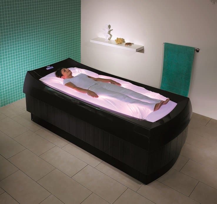 A woman lying on a futuristic LED light Therapy Bed in a Spa or wellness center, with a green tiled wall and a white shelf with decor in the background.